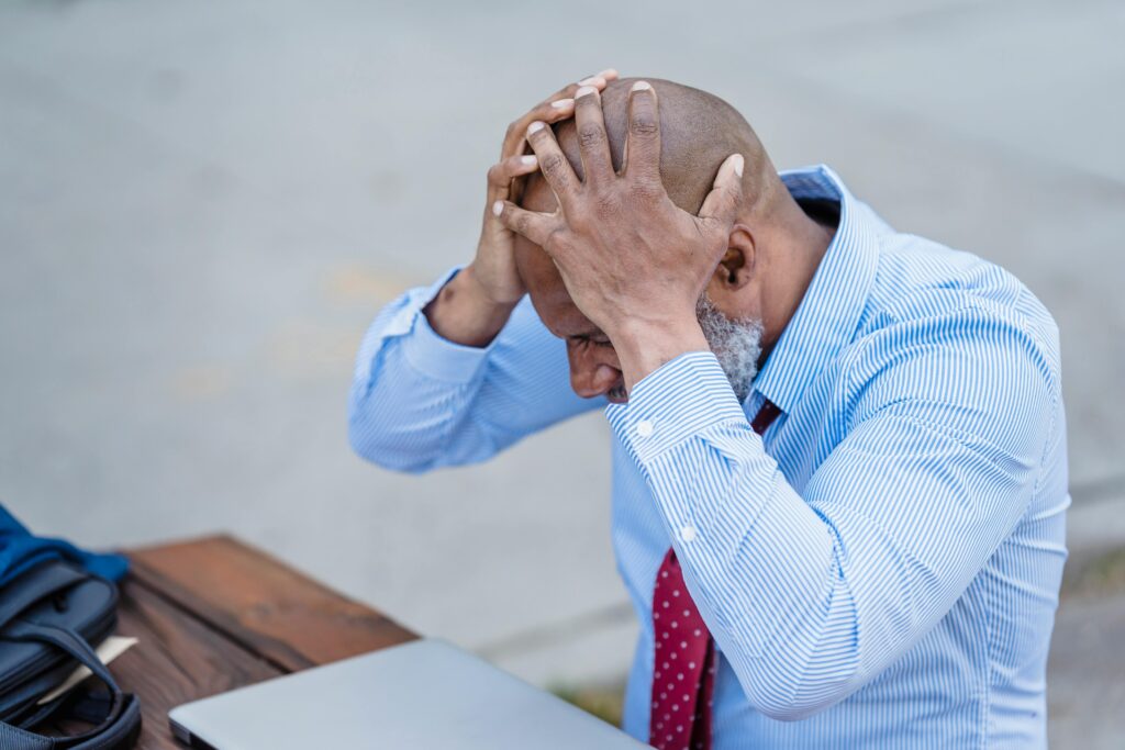 Frustrated businessman holding head in hands outdoors with laptop, corporate stress concept.